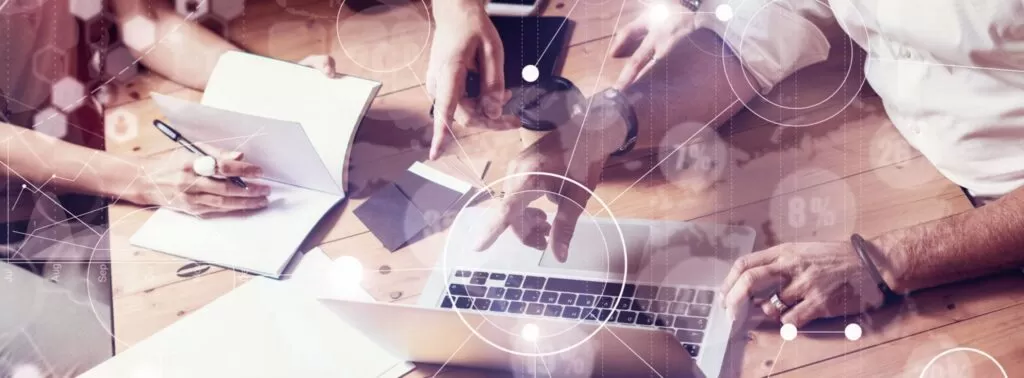 Mãos de várias pessoas trabalhando juntas em uma mesa com laptops, cadernos e papéis, representando a colaboração e o processo de análise de dados em um projeto de data analytics.