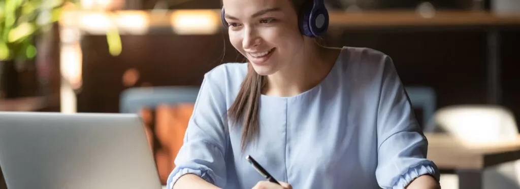 Mulher sorrindo, usando fones de ouvido e trabalhando em um laptop, representando o uso da Inteligência Artificial (IA) na educação. Conceito de aprendizado online, cursos digitais, plataformas educacionais inteligentes e o papel da IA para agilizar e otimizar o ensino e o aprendizado no mercado educacional atual.