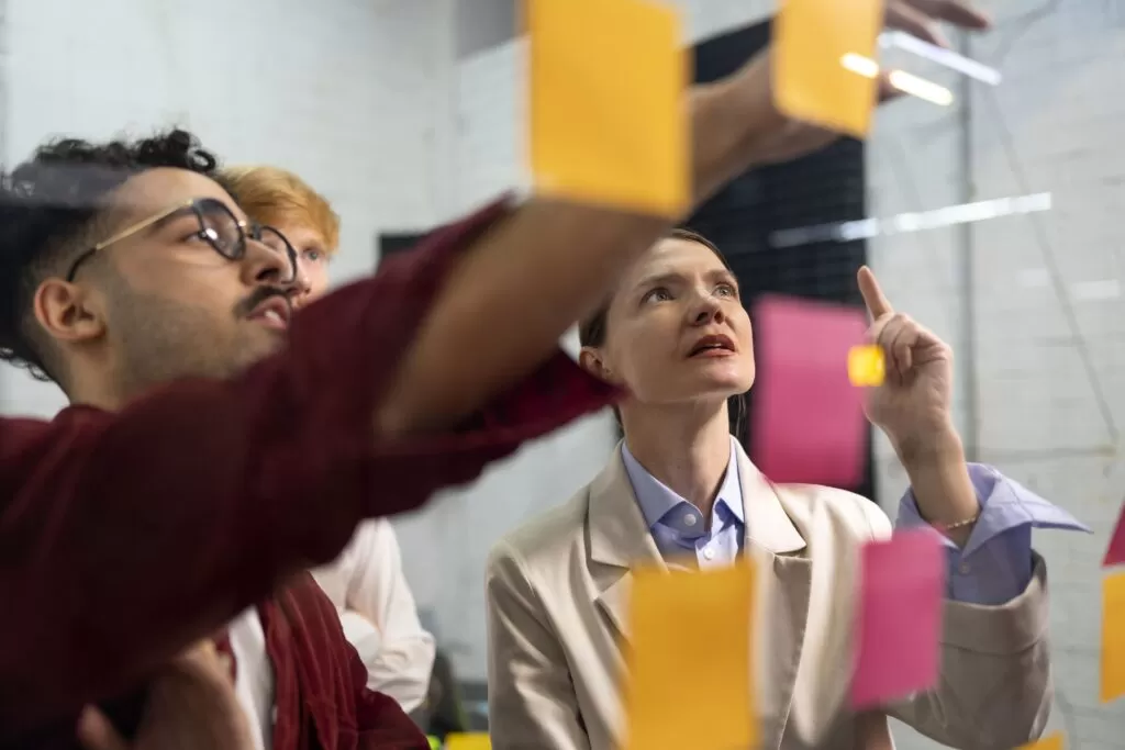 Uma equipe de pessoas trabalhando juntas em um escritório, usando post-its coloridos em uma superfície transparente para visualizar e organizar ideias, representando o processo colaborativo de geração de ideias inovadoras em uma empresa.