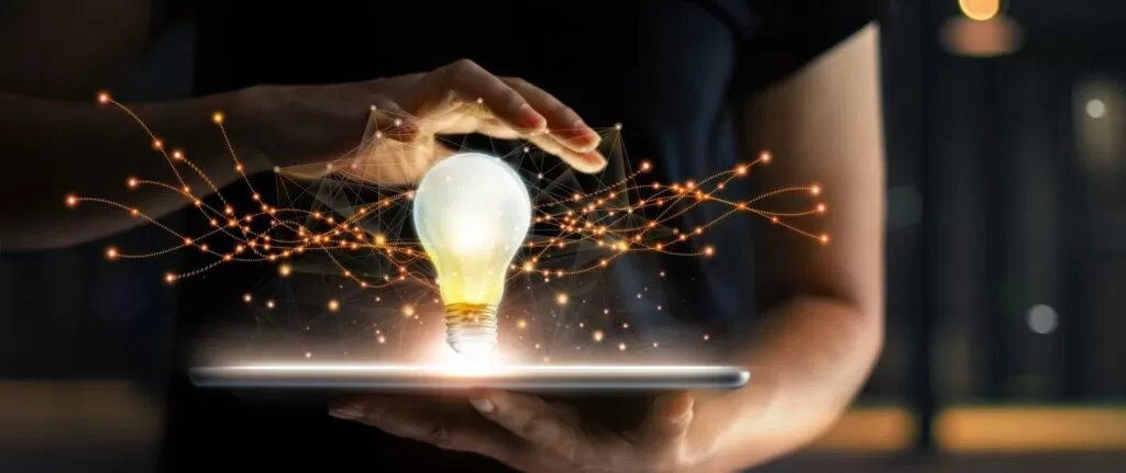 Uma pessoa segurando um tablet sobre o qual repousa uma lâmpada acesa, com linhas e pontos luminosos irradiando da lâmpada, representando a geração de ideias e o papel da tecnologia na inovação presente em nossa vida cotidiana.