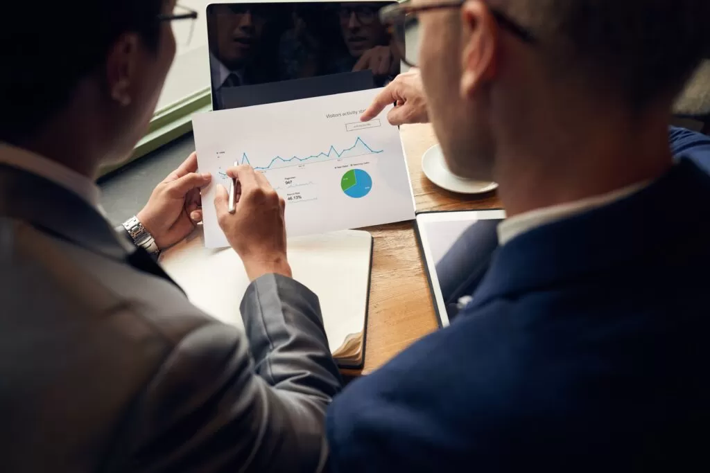 Duas pessoas trabalhando juntas sobre um documento com gráficos de tendências e participação, representando o processo colaborativo de análise que pode ser aprimorado por algoritmos de Inteligência Artificial para previsões de demanda mais assertivas.
