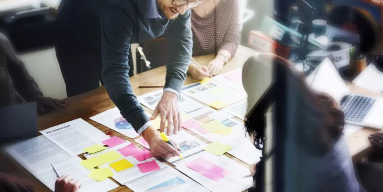 Vista superior de uma equipe de trabalho reunida em uma mesa de madeira com papéis, gráficos e notas adesivas coloridas, discutindo as etapas do planejamento de produção.