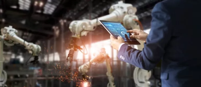 Gestor segurando um tablet com painel de dados digitais em primeiro plano, com braços robóticos industriais trabalhando ao fundo, simbolizando a tecnologia no planejamento de produção.