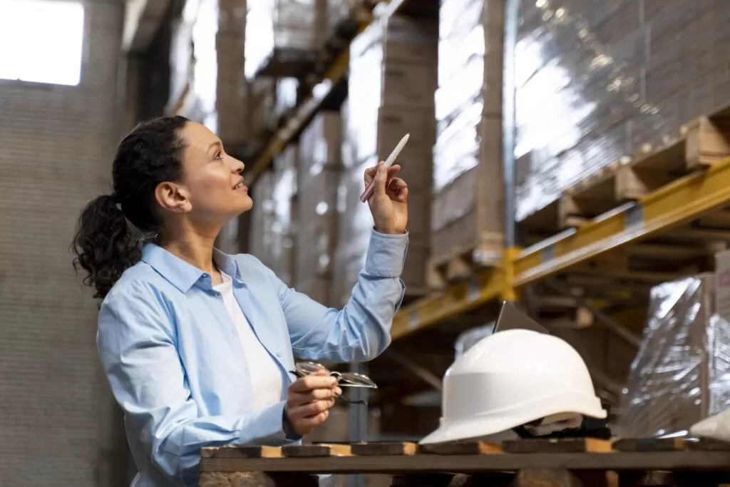 Mulher em um armazém logístico apontando para caixas em prateleiras altas, realizando a conferência de materiais necessária para o planejamento de produção.