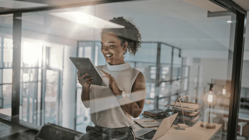 Mulher sorrindo enquanto interage com um tablet em um ambiente de escritório moderno, representando o uso da Inteligência Artificial (IA) no setor de telecomunicações.