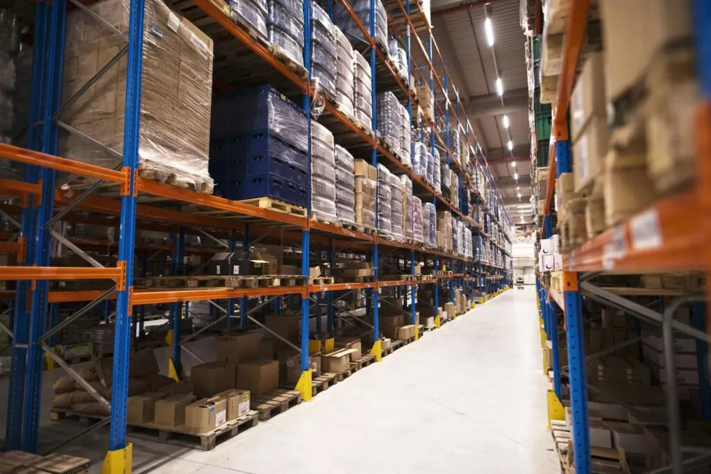 Uma visão ampla de um armazém com prateleiras altas repletas de caixas e produtos embalados, representando a escala e a complexidade do armazenamento e da logística na produção da Alpargatas, dona da marca Havaianas