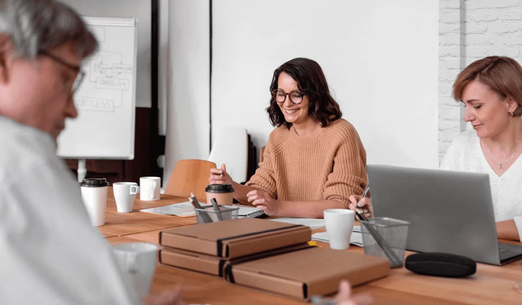Três pessoas juntas e sentadas em uma sala de reunião. Na mesa há pastas de documentos, xícaras brancas, um notebook e outros materiais de escritório. A ideia é se relacionar com o tema planejamento acadêmico.