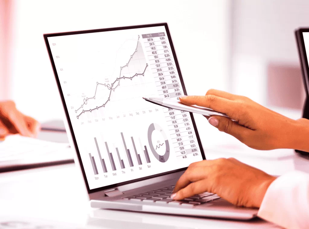 Mãos de uma pessoa apontando para uma tela de laptop exibindo gráficos de previsão de demanda com dados numéricos e linhas ascendentes, representando o Prognos, ferramenta da UniSoma que reduz erros de previsão em 30%. Foco em tecnologia para planejamento e otimização de demanda.