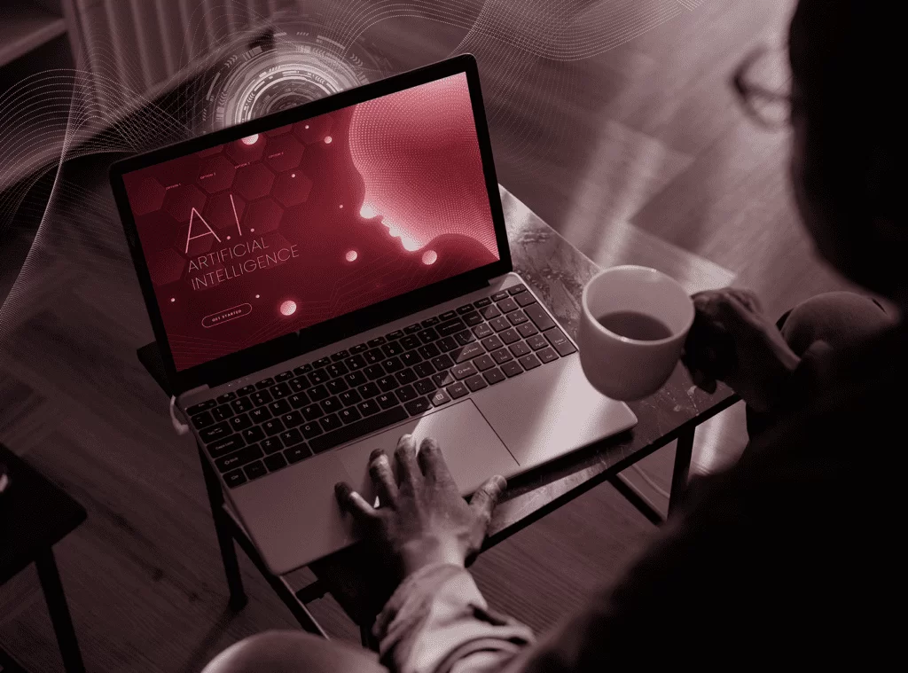 Pessoa sentada em frente a um notebook segurando uma xícara branca com café. Na tela do notebook está escrito "A.I", o que se relaciona com o impacto da inteligência artificial no planejamento de pricing