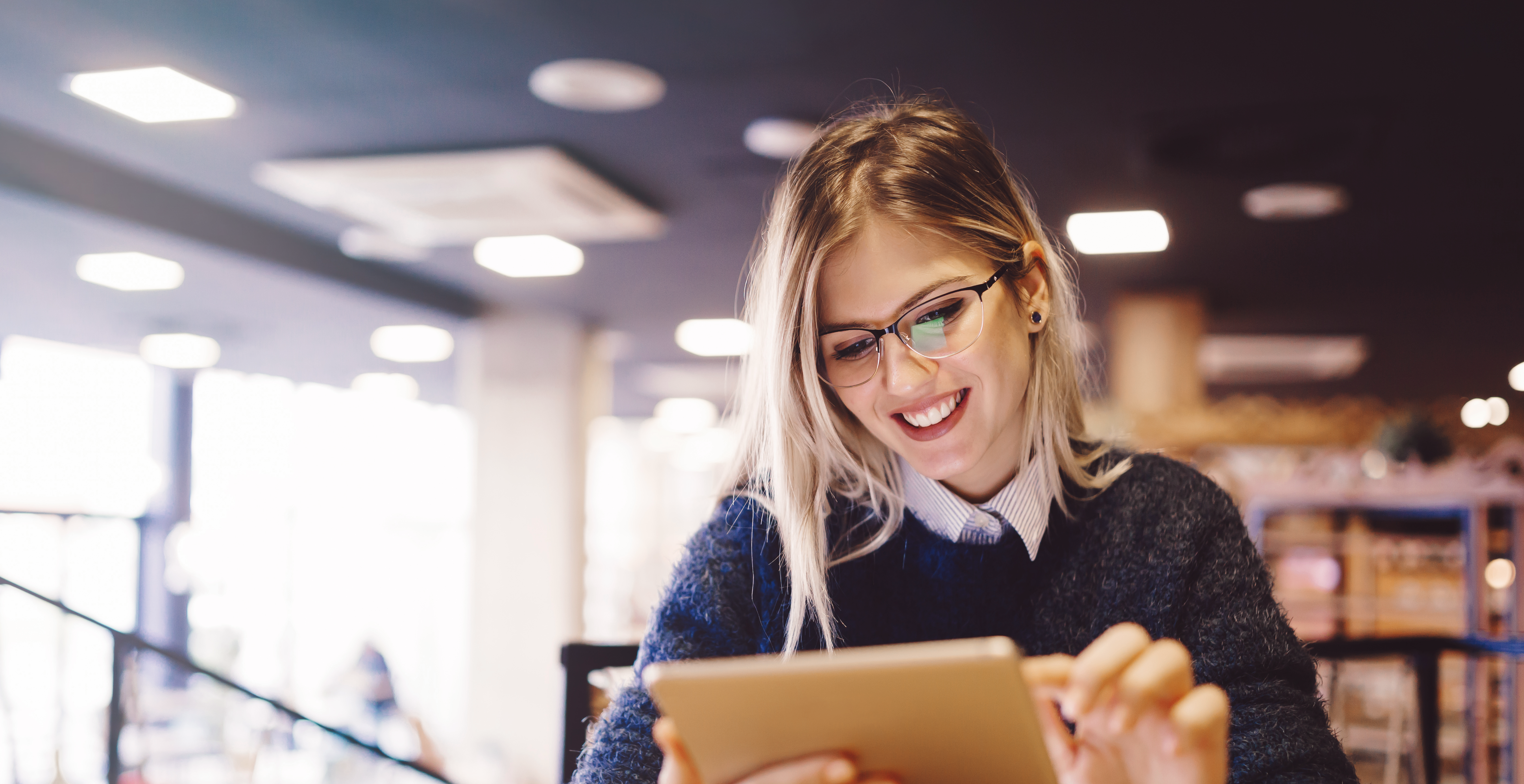 Mulher sorrindo enquanto usa um tablet em um café moderno, simbolizando organização e produtividade. A imagem reflete o tema do artigo 'O que é planejamento acadêmico e como desenvolvê-lo'