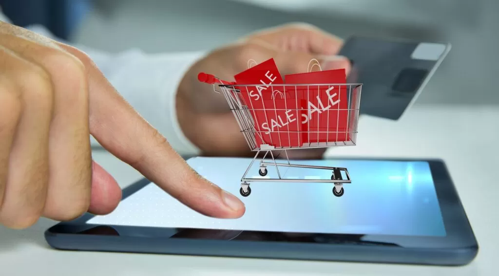 Importância do Pricing e Inteligência de Mercado. | UniSoma Mão utilizando tablet para compras online com carrinho de supermercado virtual e etiquetas de oferta, representando estratégias de pricing e inteligência de mercado.