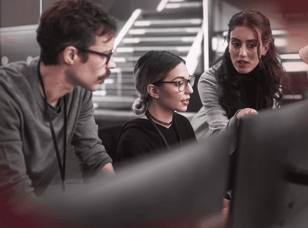 Três profissionais lado a lado, sendo um homem e duas mulheres, em um ambiente corporativo. Essas pessoas estão conversando entre si em frente a monitores. Representa a uso de tecnologia para analisar a evasão de clientes.