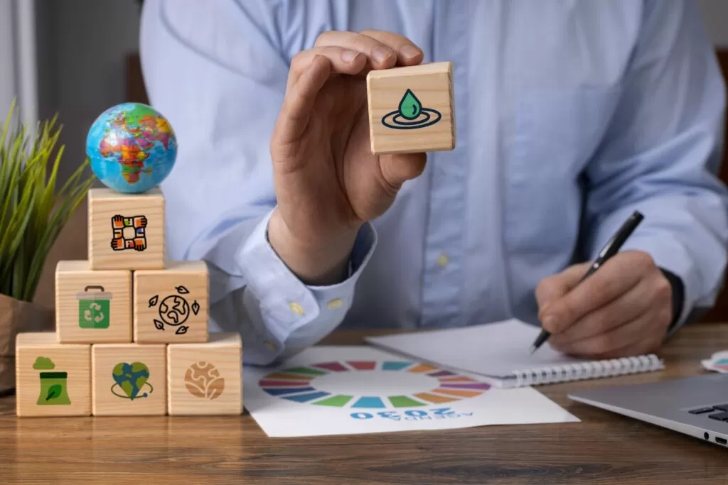 Pessoa apresenta cubo de madeira com gota d'água, simbolizando sustentabilidade (ESG). Ao redor, outros cubos com ícones de meio ambiente, sociedade e governança, destacando a importância do ESG para empresas.