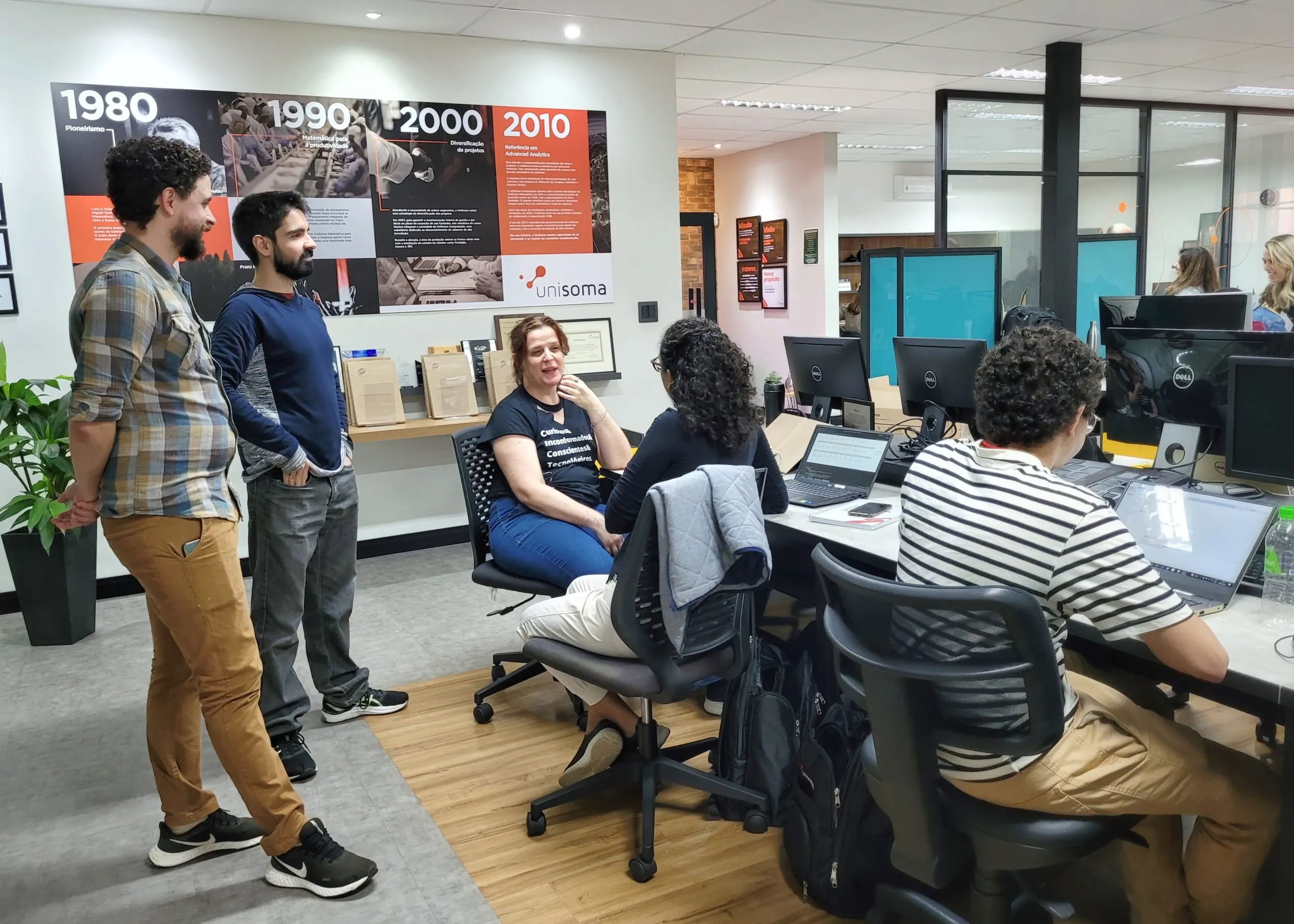 Profissionais da UniSoma conversando no escritório, com o mural da história da empresa ao fundo